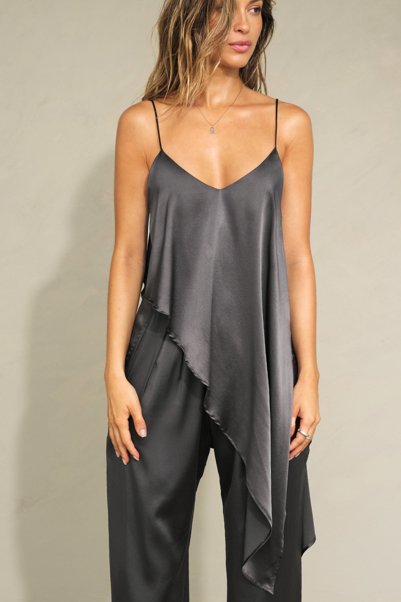 Front view of model posing the flowy iron Asymmetrical Silk Tank with a V-neckline, asymmetrical hem and an open back with thin, versatile straps that can tie straight or crisscross at the back