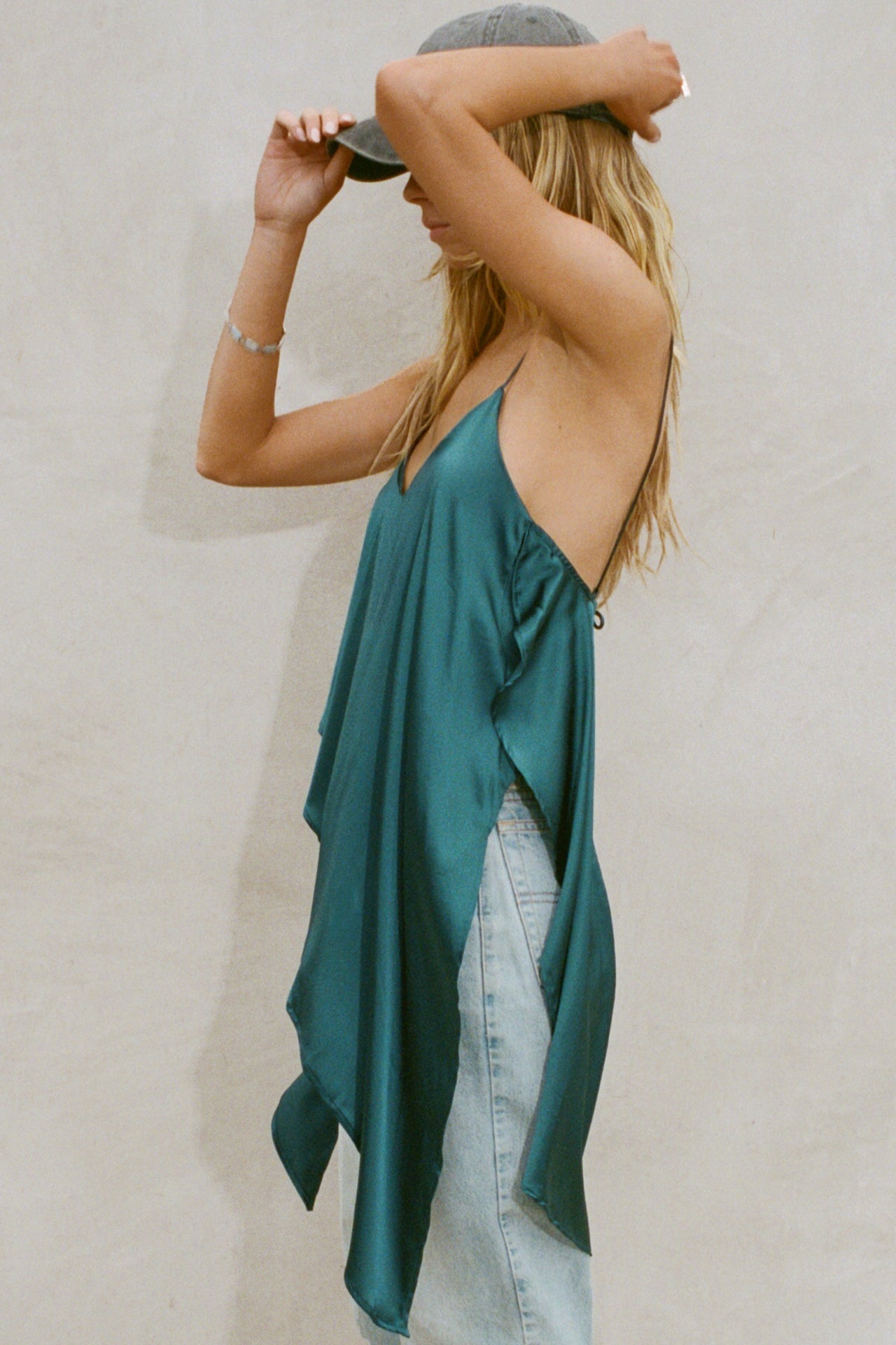 Side view of model posing the flowy lagoon Asymmetrical Silk Tank with a V-neckline, asymmetrical hem and an open back with thin, versatile straps that can tie straight or crisscross at the back