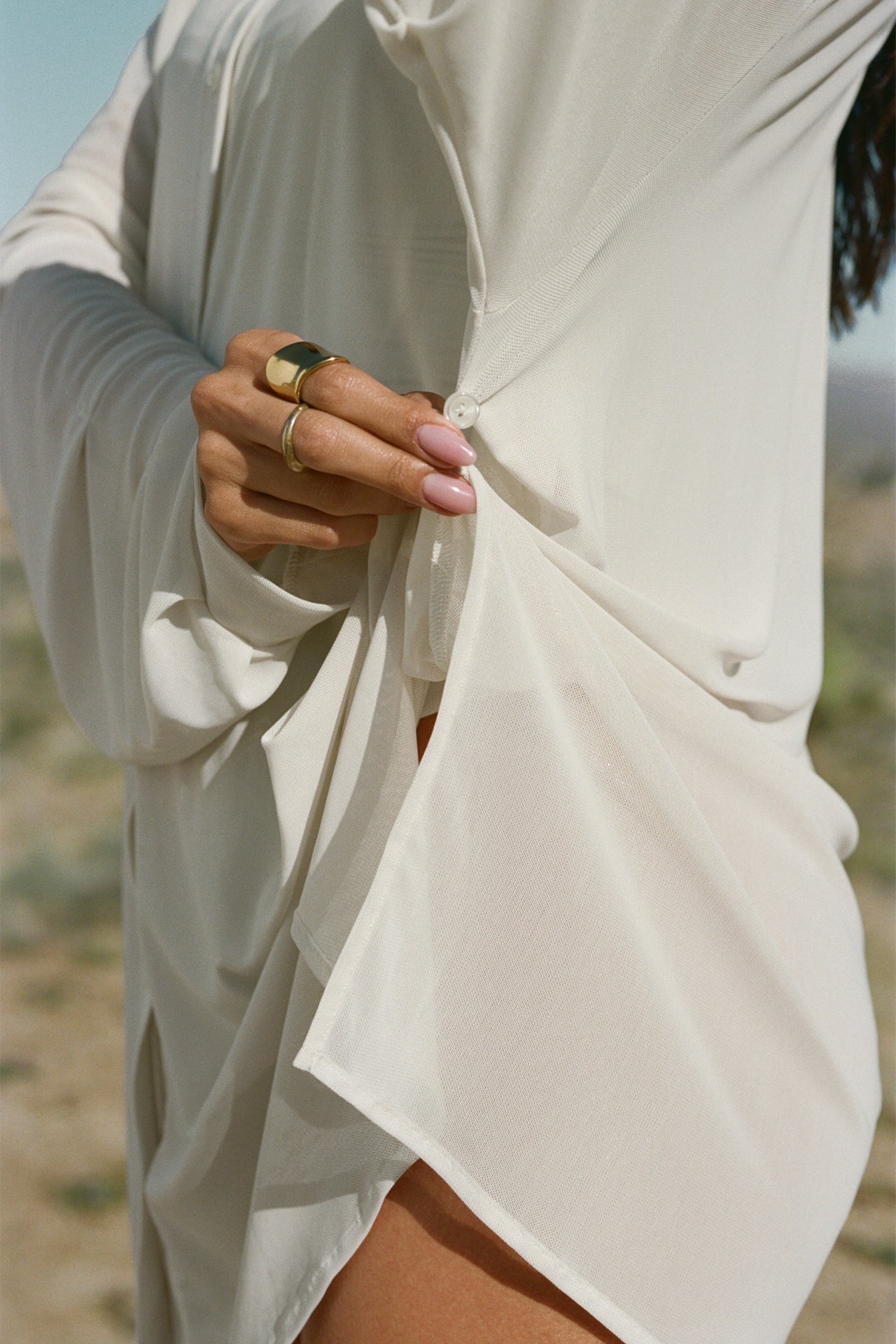 Close up detail side view of model posing in the sheer, lightweight porcelain  mesh Oversized Sheer Button Down long sleeve shirt with a classic collar, front button closures, dropped sleeves and buttons below the underarms for adjustable cinching
