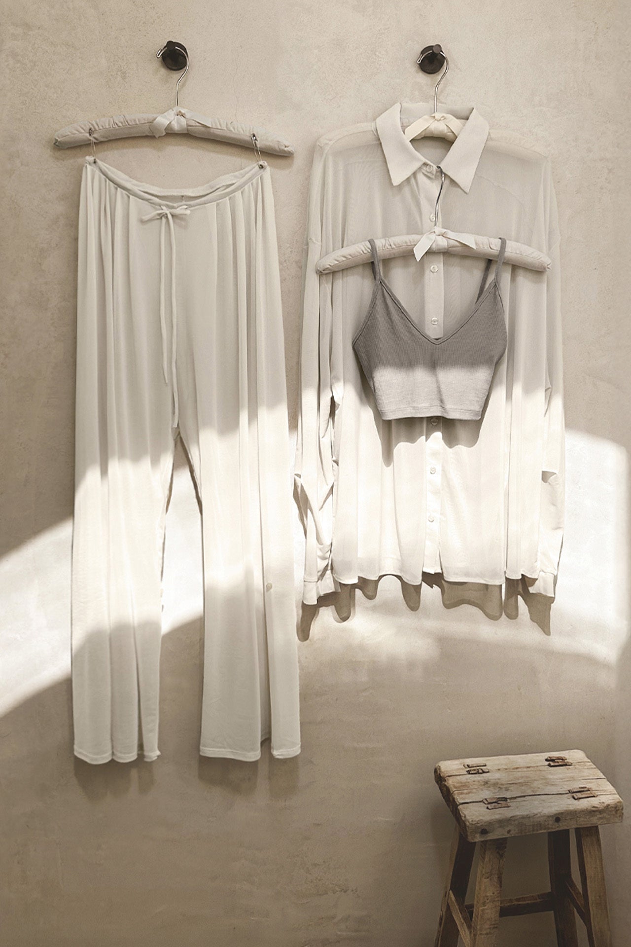 Front view of the porcelain mesh Oversized Sheer Button Down, the porcelain mesh Pleated Sheer Pant and the platinum flexrib Strappy Crop Tank hanging up on hangers in a room with a stool